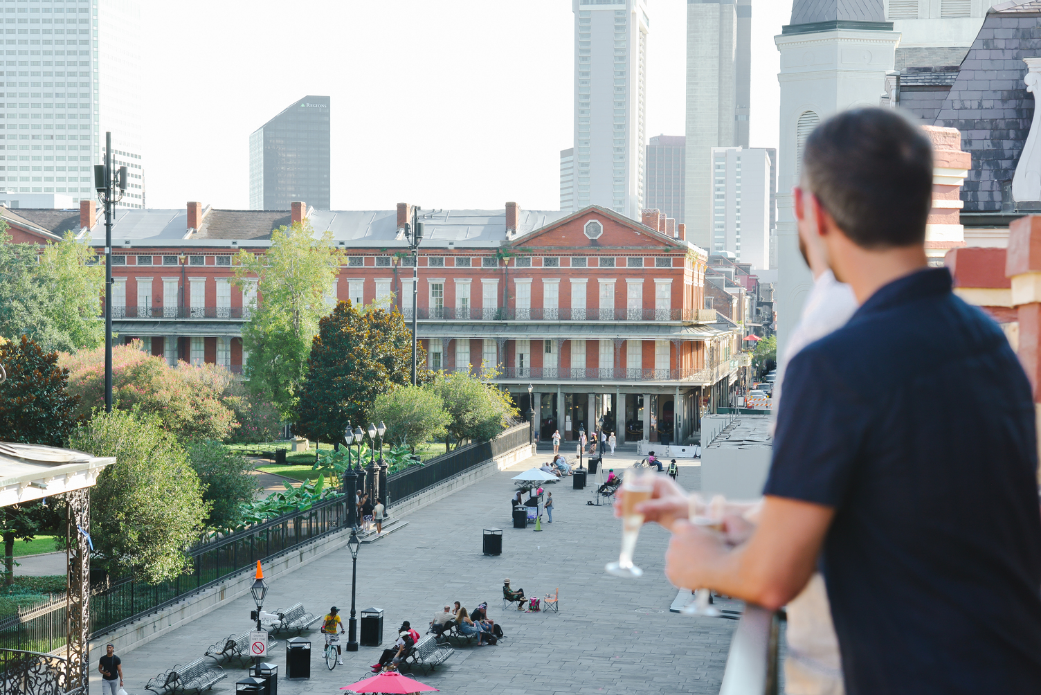 A man stands on a balcony overlooking a city square with historic buildings and a park.